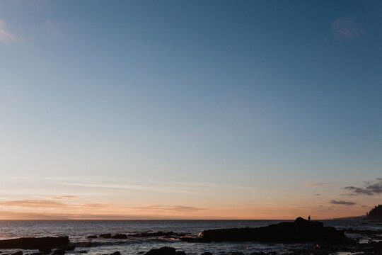 A faraway man stands looking to the ocean at a west coast sunrise.