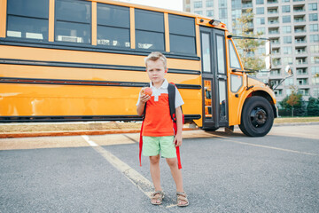 Caucasian boy student with backpack near yellow bus on first September day. Child kid eating apple...
