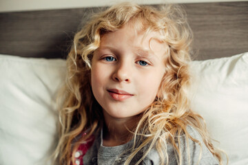 Pretty beautiful Caucasian blonde girl with long messy hair and blue eyes sitting on bed at home...