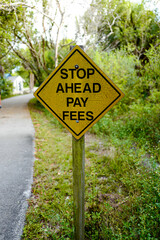 Rusty yellow sign erected on a tropical pedestrian and bicycle trail to warn users to stop ahead and pay fees. Vertical layout.