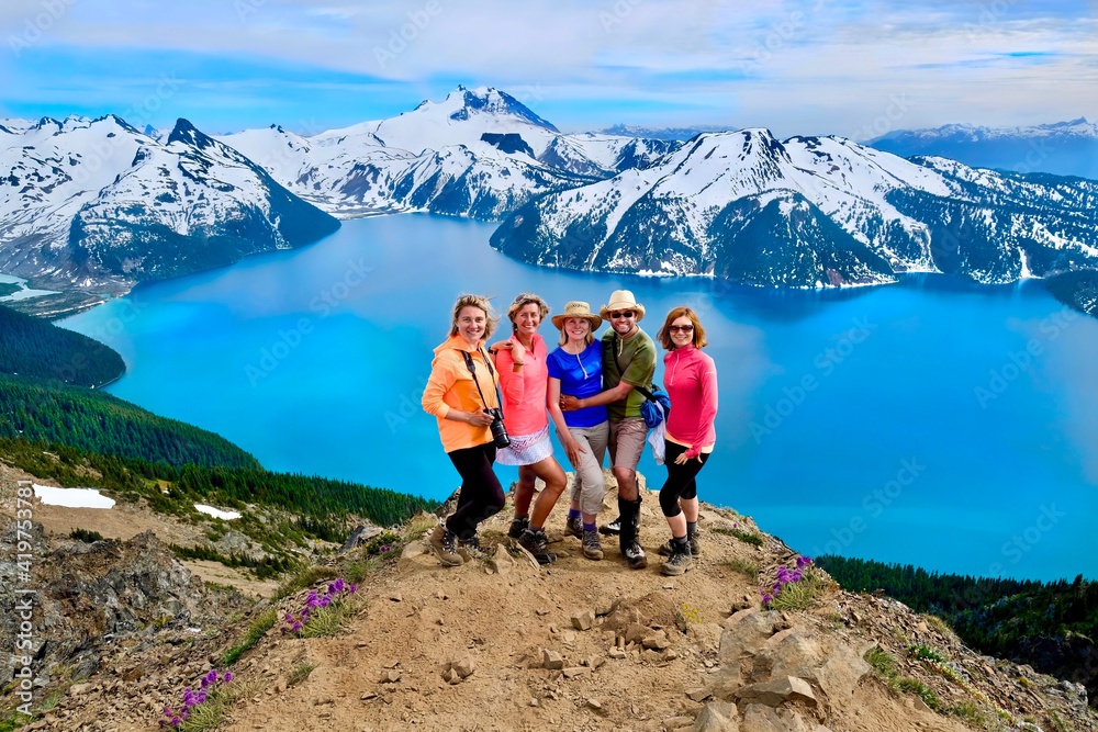 Wall mural cheerful group of people on cliff above turquoise lake snowcapped mountains. garibaldi lake from pan