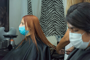 Two clients cutting their hair in a hairdresser with a protective mask with social distancing against covid-19. Caucasian lady with red hair at the stylist.