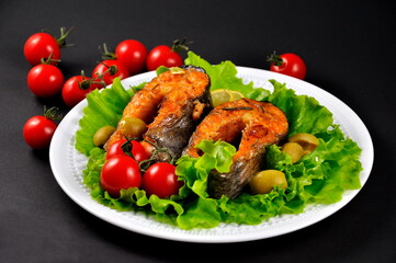 Slices of baked trout on a white plate, with lemon and cherry tomatoes on a black background.