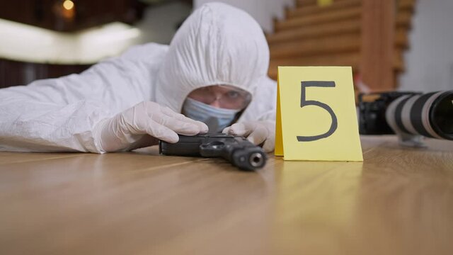 Weapon lying on floor with blurred forensic scientist examining evidence. Focused Caucasian woman working at crime scene indoors. Murder investigation and forensic science concept.