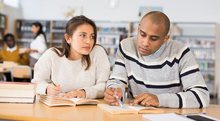 Positive hispanic man and woman spending time together in library, talking, reading books and making notes