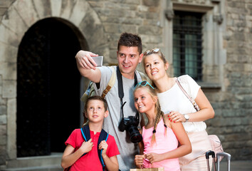 Young glad tourist family of four strolling on city streets and taking selfie