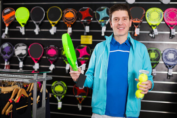 Sporty smiling male in uniform is holding new rocket and balls for padel in the store.