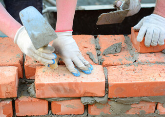 Two bricklayers are laying bricks of a brick wall, foundation using a mason's trowel and mortar. A close-up of a brickwork from burnt clay bricks.