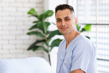 middle age caucasian short black hair happy male patient wear blue hospital shirt sitting on bed looking at camera smiling alone in ward room with blurred green tree and white brick wall background