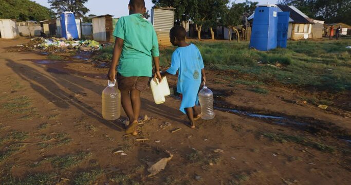 Water crisis. Rear close-up view of two young black African boys carrying home dirty unsafe drinking water for domestic use.  Poor living conditions and no access to clean running wate