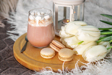 Fototapeta na wymiar A glass of hot cocoa with marshmallows, macaroni cakes and white tulips on a tray in bed close up. Cozy spring home concept
