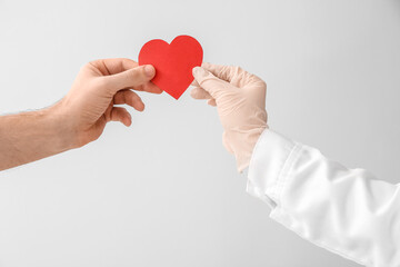Hands of cardiologist and patient with red heart on light background