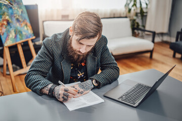 Fototapeta na wymiar Stylish freelancer doing his remote work at home. Professional portrait of a hipster person sitting at table with a laptop and smartphone.