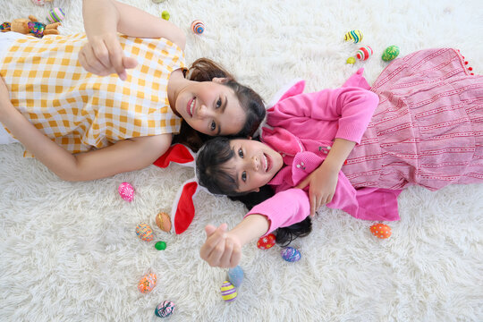The young mother and her daughter lay down together on the carpet to playing easter eggs. They showing mini heart sign their hands.