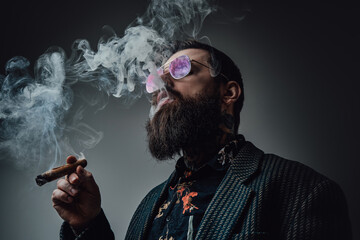 Elegant man in retro style poses in dark background smoking cigar. Portrait of a bearded guy with stylish coiffure.