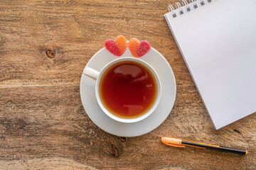 Tea break concept. Tea cup, marmalades in the form of heart, blank notebook paper, pen on wooden table. Top view.