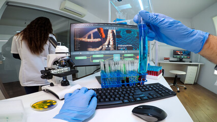 FPV of doctor chemist holding a flask of blue solution looking at it and typing on computer medical results. Scientists examining vaccine evolution in medical lab using high tech chemistry tools
