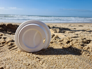 Single-use white plastic cup lid cover on the summer sandy beach in front of the water ocean. Dirty...