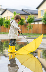 Boy holds yellow umbrella standing in puddle outside. On sunny day happy kid walks near the house, plays, has fun