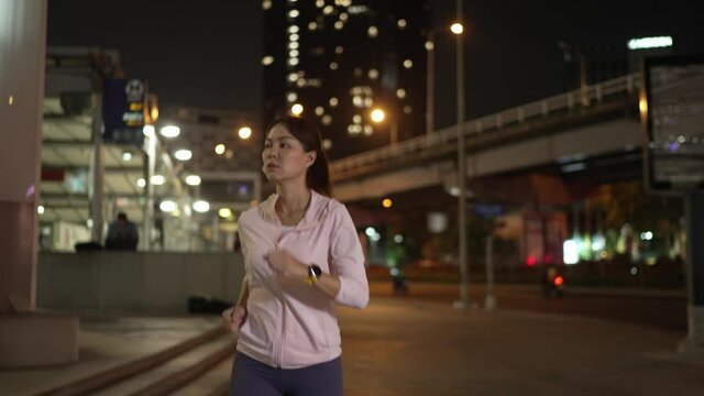 Slow-motion,Font Follow Camera View.Female athlete in hooded shirt Jogging at night City streets with lots of lights in the background.
