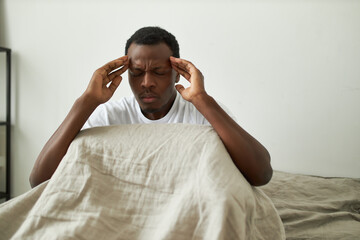 Unhappy young African man sitting on bed closing eyes and massaging temples to soothe pain,...