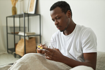 Sick unhealthy young African American man in white tee staying in bed because of flu, having high temperature, holding pills and glass of water, taking antiviral medicine. Treatment and sickness