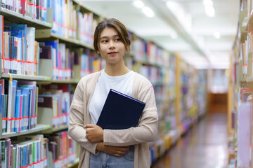 Portrait of Smart Asian woman university student reading book and looking at camera between bookshelves in campus library with copyspace..