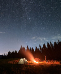 Hiker standing near campfire under night starry sky.