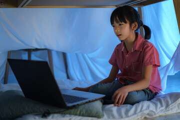 Asian girl child to make a camp to play imaginatively watching a film on laptop in the darkness of the camp in living room at home. - Powered by Adobe