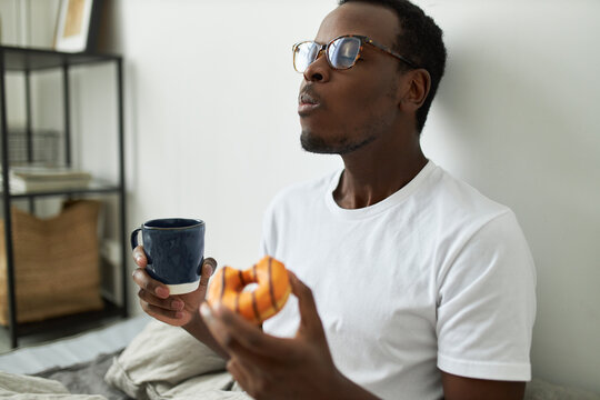 Indoor shot of stylish hungry young dark skinned man in glasses eating glazed orange doughnut with great relish, closing eyes with pleasure, enjoying every bite of sweet dessert, drinking morning tea