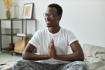 Portrait of handsome joyful young dark skinned man in comfortable clothes seated on bed with hands pressed together, making namaste as greeting sign, feeling grateful for everything, smiling