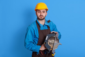 Pleasant looking handsome worker holding tool in both hands, looking directly at camera with serious expression, wearing brown apron, blue overall and yellow helmet, against color wall.