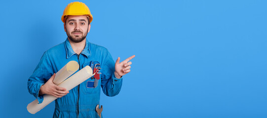 Male worker holding blueprints and pointing aside with index finger, unshaven engineer wearing yellow helmet and blue uniform looking at camera, copy space for advertisement and promotional text.