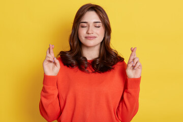 Pretty superstitious woman crosses fingers, prays for better, looks calm and relaxing, closes eyes, wears orange jumper, poses indoor over bright yellow background, anticipates important results.