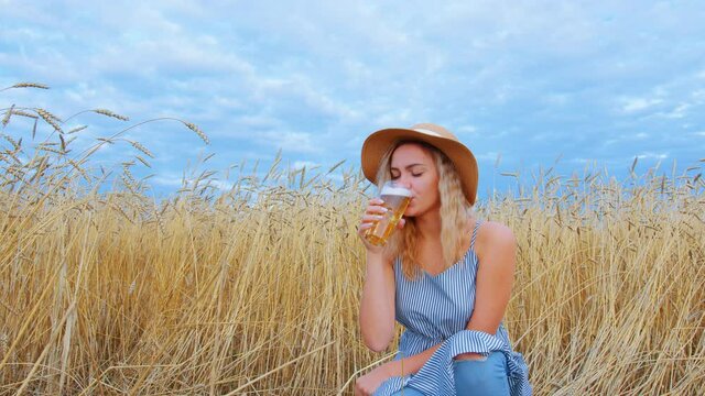 Young beautiful woman drinks beer from a glass in a cereal field. A woman in a blue dress and hat drinks alcohol on the background of nature and the sky