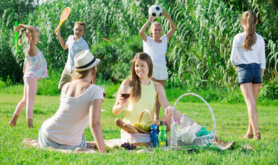 Portrait of young mums on picnic in summer park with happy children playing behind..