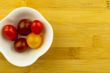 Small colorful tomatoes in a white bowl on a wooden chopping board. Artificial light from two lamps, soft shadows