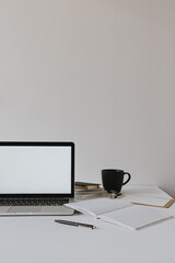 Minimalist home office desk workspace. Laptop with blank copy space mockup on table with coffee cup, paper sheet, stationery against white wall. Work, business background.