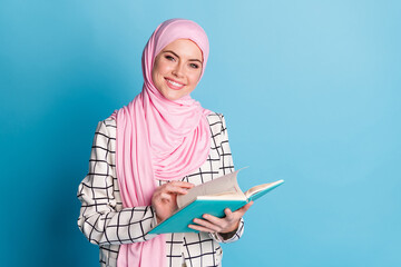 Portrait of attractive cheerful muslimah girl reading novel book isolated over vibrant blue color...