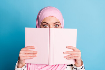 Close-up portrait of funny scared amazed muslimah girl closing face book isolated over vibrant blue...