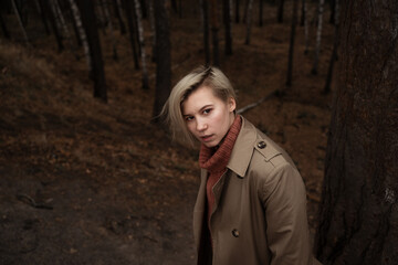 a girl of non-binary appearance with short hair stands in the park in a trench coat, spoils