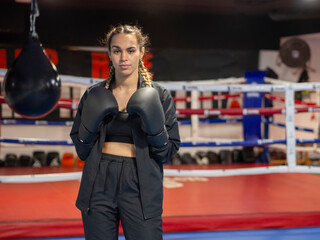 Boxer girl with black boxing gloves standing in front of a boxing ring and looking at camera. Young woman in sportswear working out at a gym. Kickboxing training for healthy lifestyle. Copy space