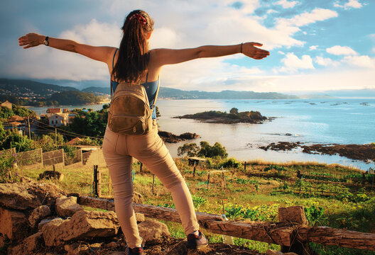 Chica con mochila de pi&eacute; sobre un muro y los brazos extendidos. Delante tiene fincas y la r&iacute;a de Pontevedra. Se pueden ver alguna casas y la costa de Mar&iacute;n. En la r&iacute;a se ve la isla de "O Santo do Mar"