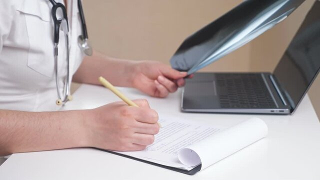 nrecognizable female doctor in white robe works with X-ray picture and papers making notes on clipboard, sitting at table with modern laptop in office closeup