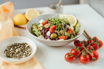 Fresh salad of vegetables, cucumber, tomato, spinach, arugula and olives and cheese in a plate on a white table, pumpkin and sunflower seeds with yellow lemon on wodden board. Food background, recipe.