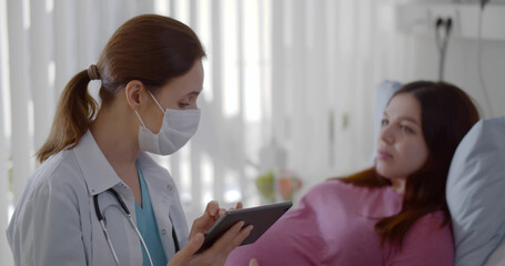 Obraz premium Female doctor in safety mask discussing with pregnant woman over digital tablet in hospital