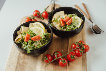 Two black bowl of rigatoni pasta with arugula, green peas, basil and cherry tomato served on wooden board. Healthy food concept, side view. Italian pasta recipe