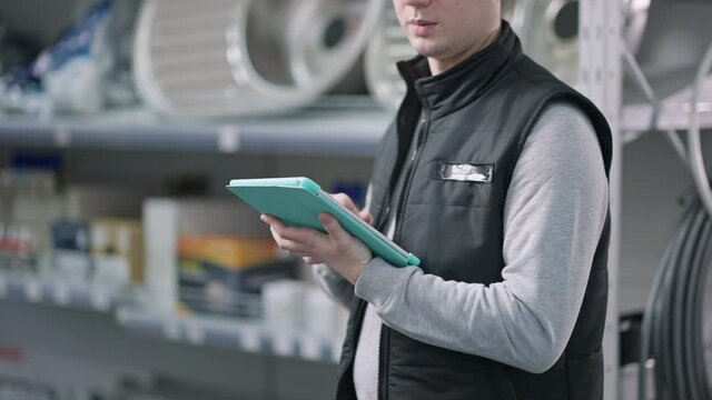 Unrecognizable Caucasian man swiping tablet and looking around on shelves in hardware store. Focused young male employee working in building supplies shop. Service and industry.