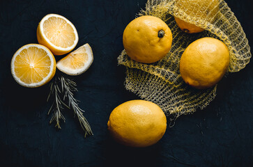 Lemons spilling out of a yellow net bag along with cut lemons, on black textured background