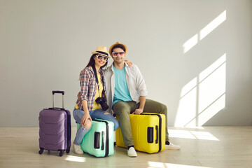 Fototapeta na wymiar Happy couple travelers in sunglasses ready for vacation travel sitting on luggage bag and hugging. Joyful young tourists posing for camera studio shot portrait with copy space. Tourism, holiday trip